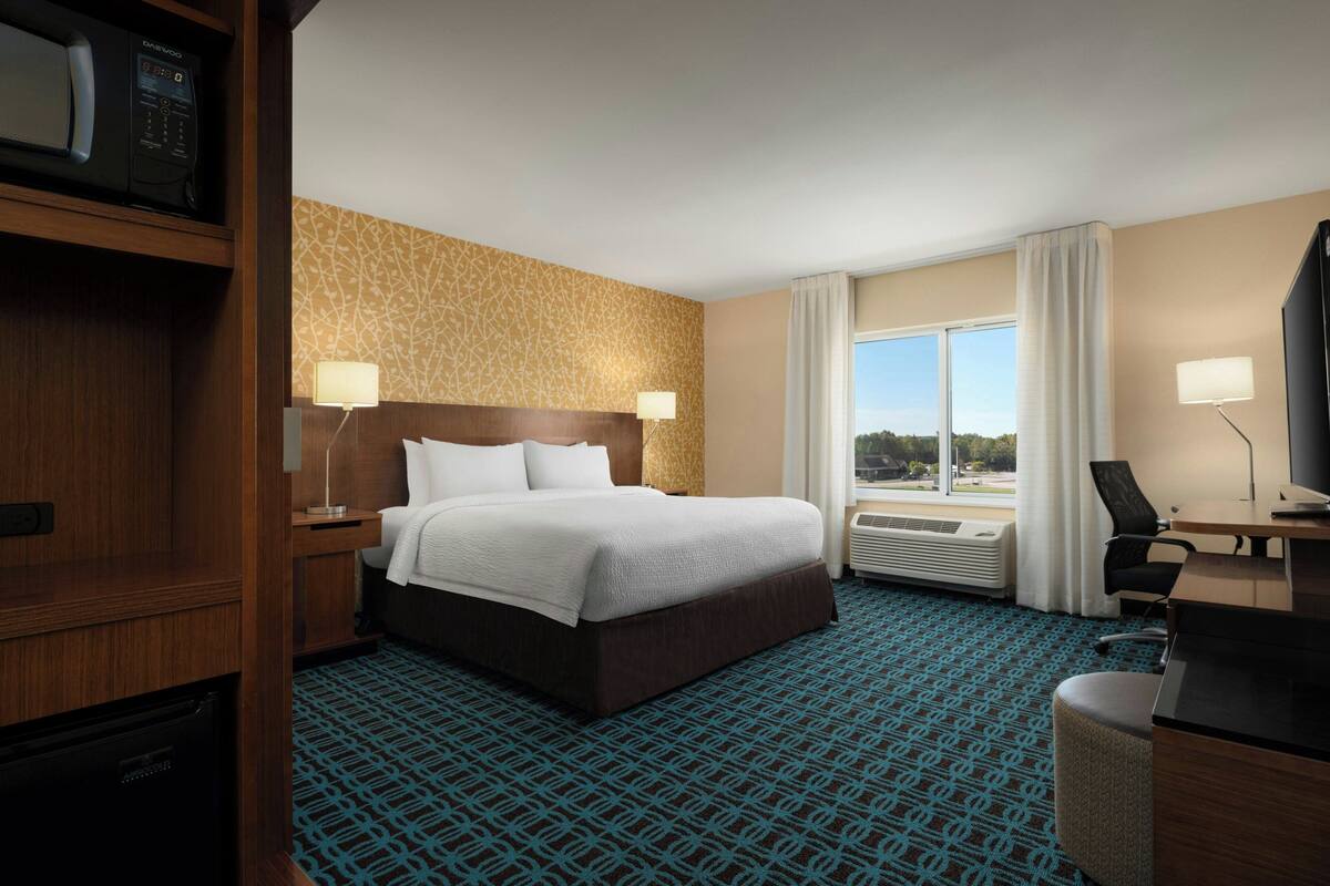 A hotel room with a large bed, two bedside lamps, a desk with a chair, and a view of the outdoors through a window.