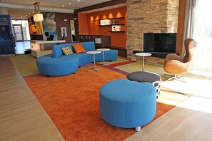 A modern lounge area with a blue sofa, ottoman, and a curved chair. There is a fireplace and a reception desk in the background. - Fairfield Inn & Suites by Marriott London (London)