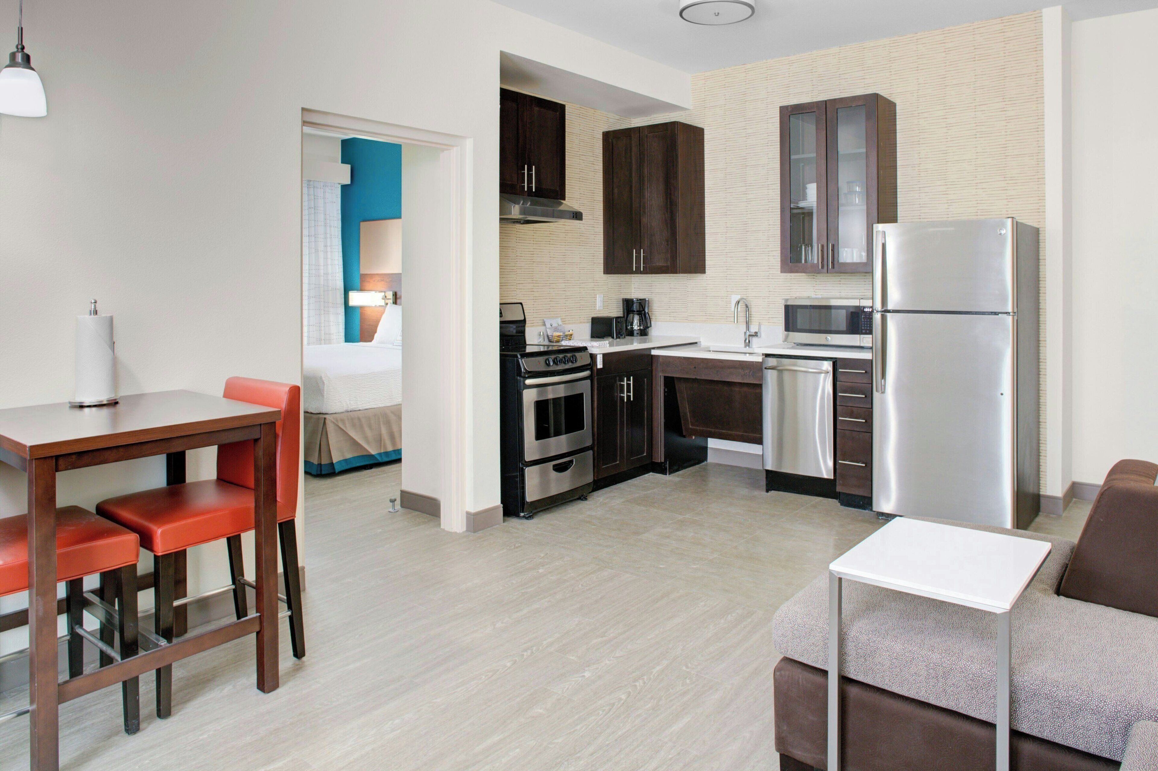 A modern kitchen with dark cabinets, a stainless steel refrigerator, and a microwave. There is a dining area with a table and chairs.