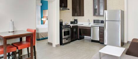 A modern kitchen with dark cabinets, a stainless steel refrigerator, and a microwave. There is a dining area with a table and chairs.