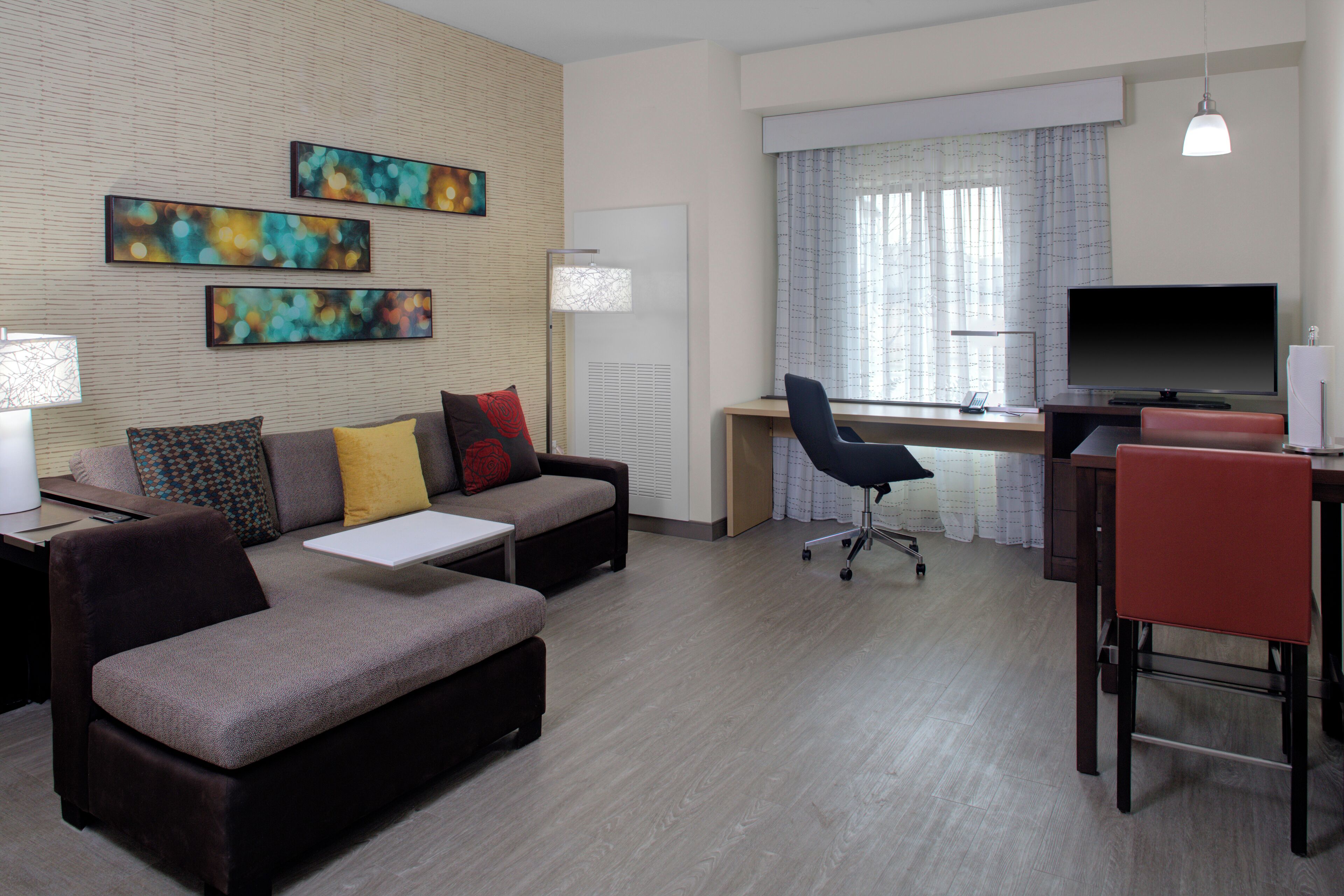 Et moderne hotelværelse med sofa, skrivebord, fjernsyn og kunst på væggen.