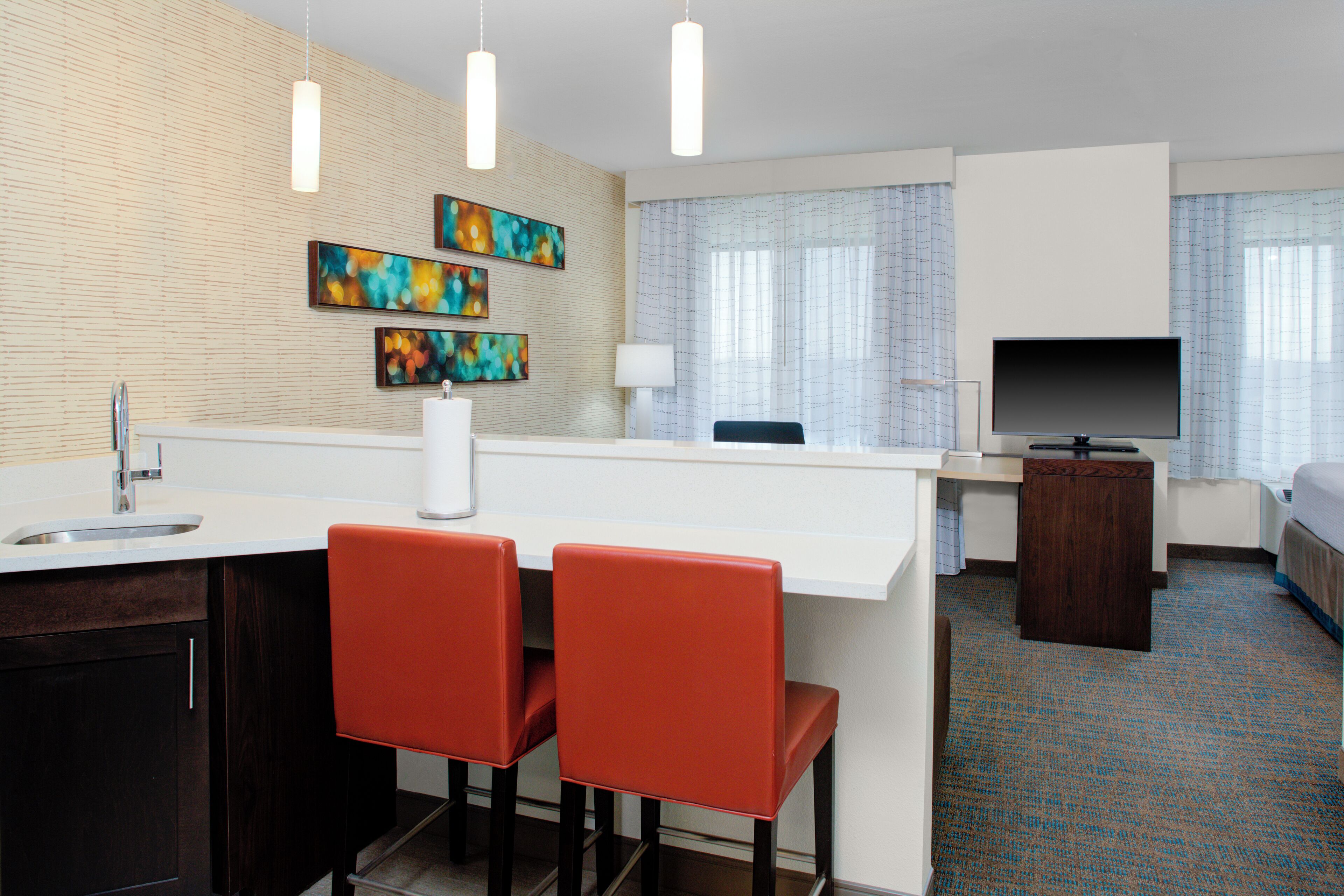 Quarto de hotel moderno com cozinha compacta, área de refeições com duas cadeiras vermelhas, televisão de tela plana e obras de arte na parede.