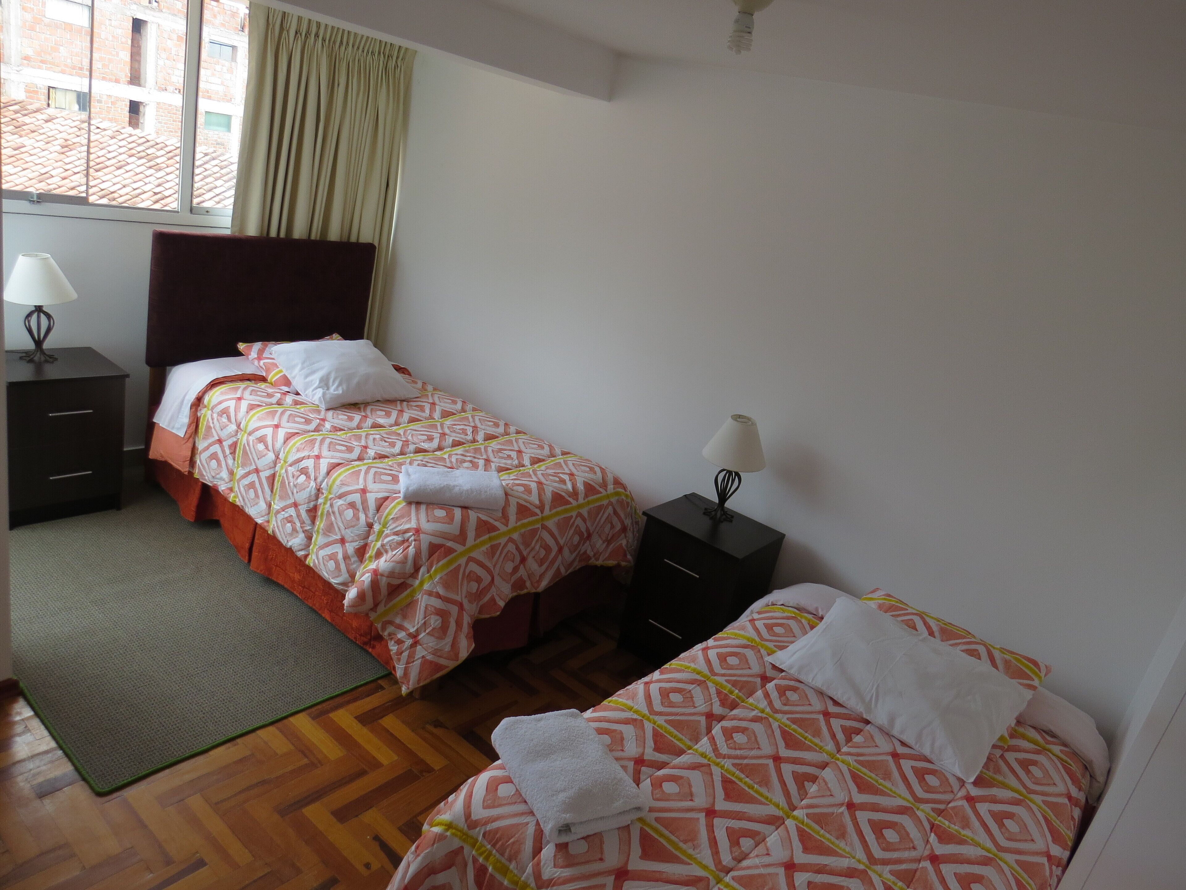 Habitación de hotel con dos camas, una ventana con cortinas, una mesita de noche con lámpara y una alfombra pequeña.