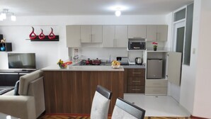 A modern kitchen with white cabinets, a microwave, and a refrigerator. There is a dining area with a sofa and chairs. - Apartamentos Quewe (Cusco)