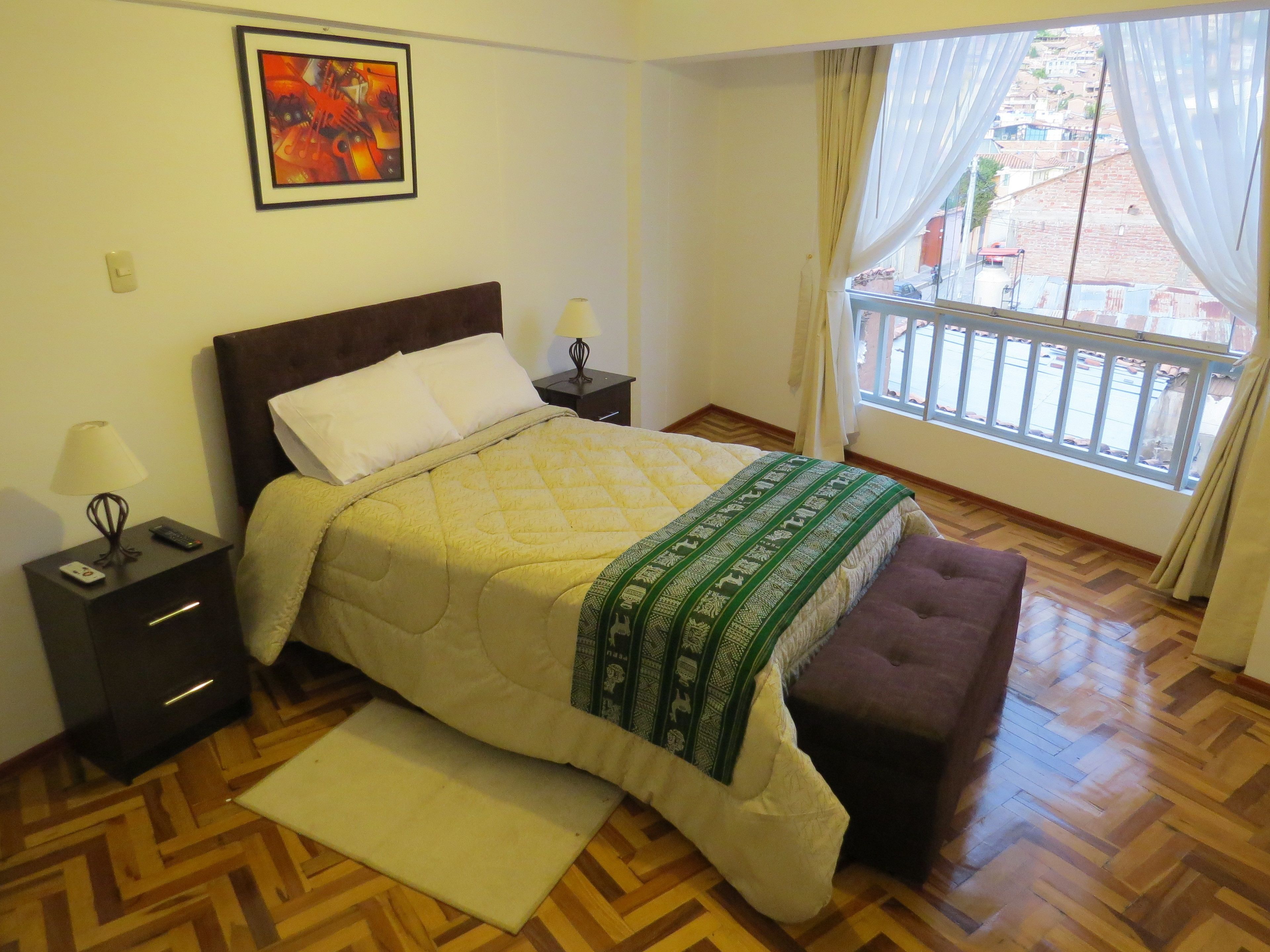A bedroom with a bed, a nightstand, a lamp, a window with curtains, and a view of buildings outside.