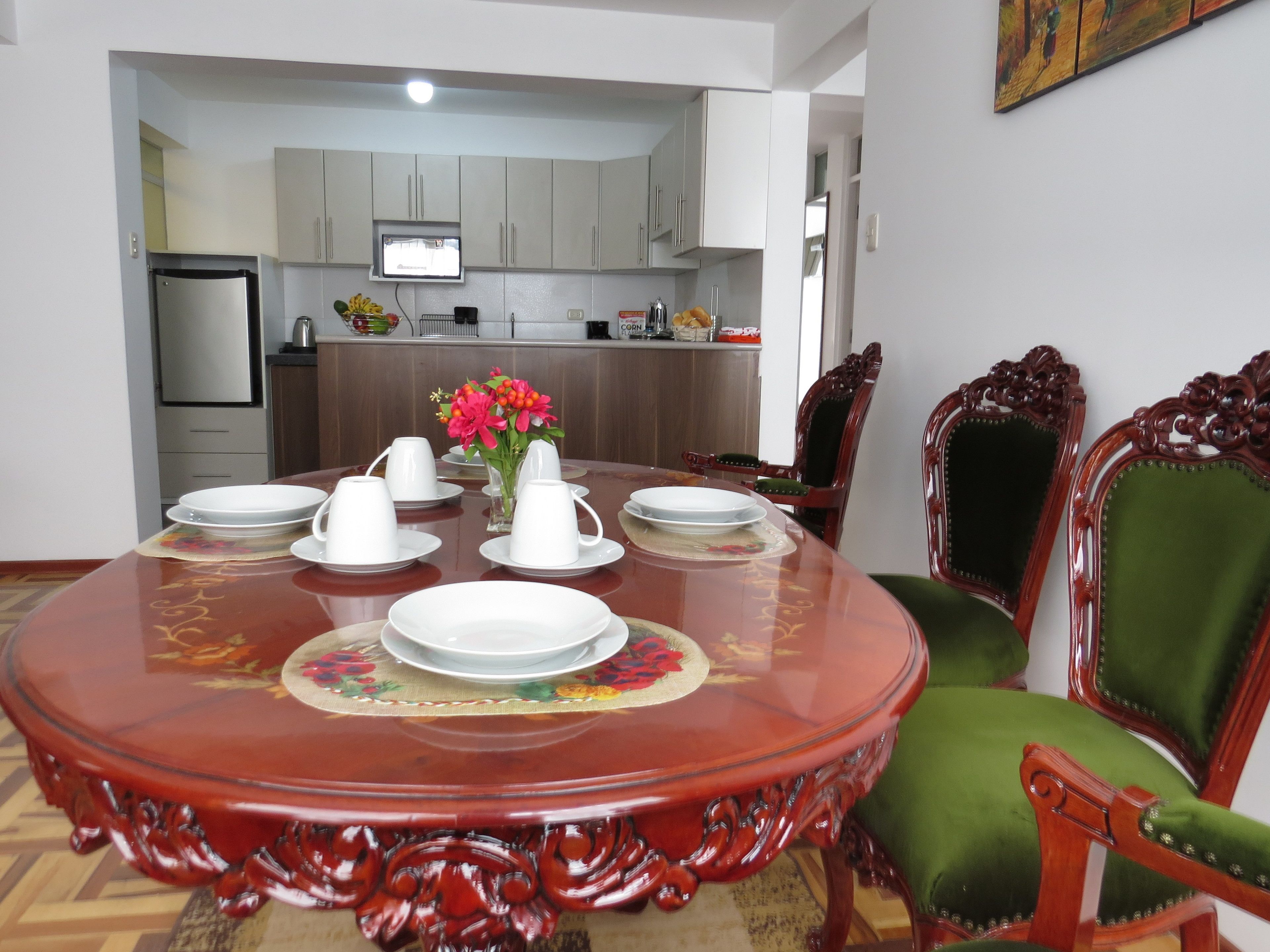 Comedor con una mesa redonda dispuesta para seis personas, rodeada de sillas ornamentales. Se observa una cocina al fondo.