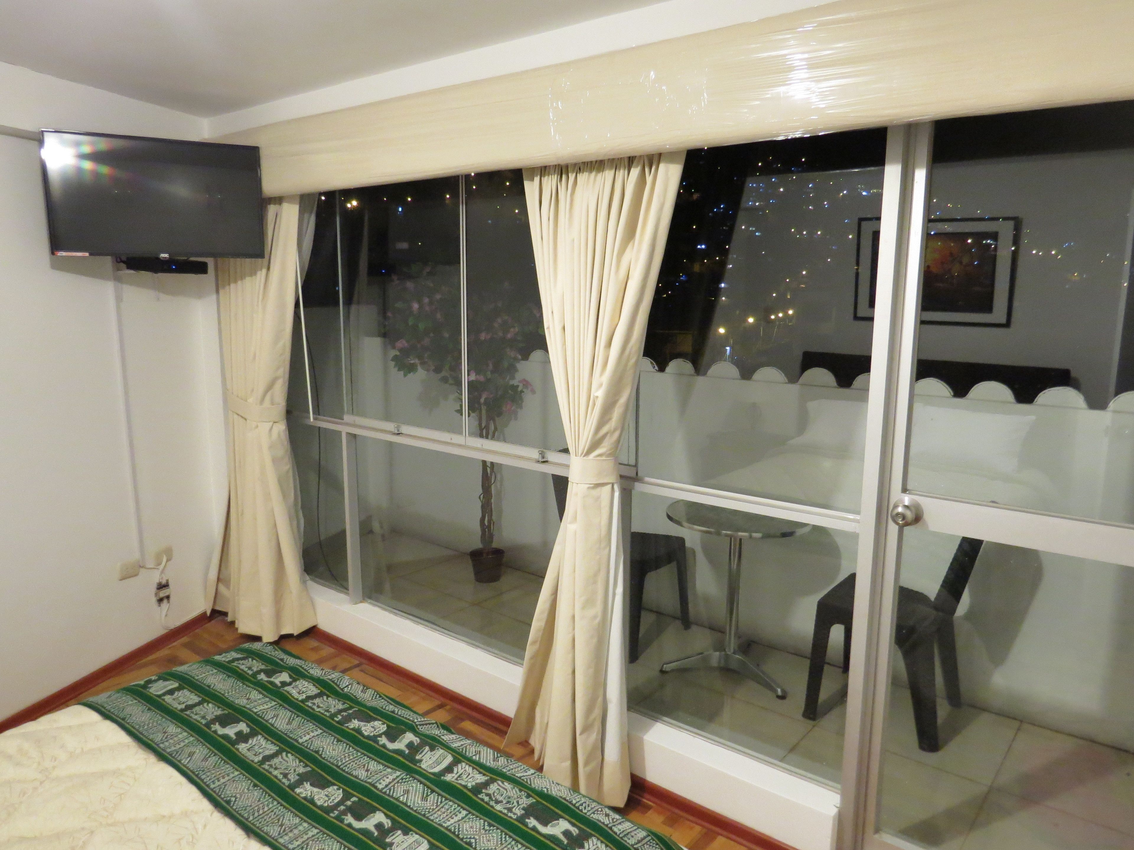 A room with a bed, a TV mounted on the wall, a window with curtains, a small table, and a chair.