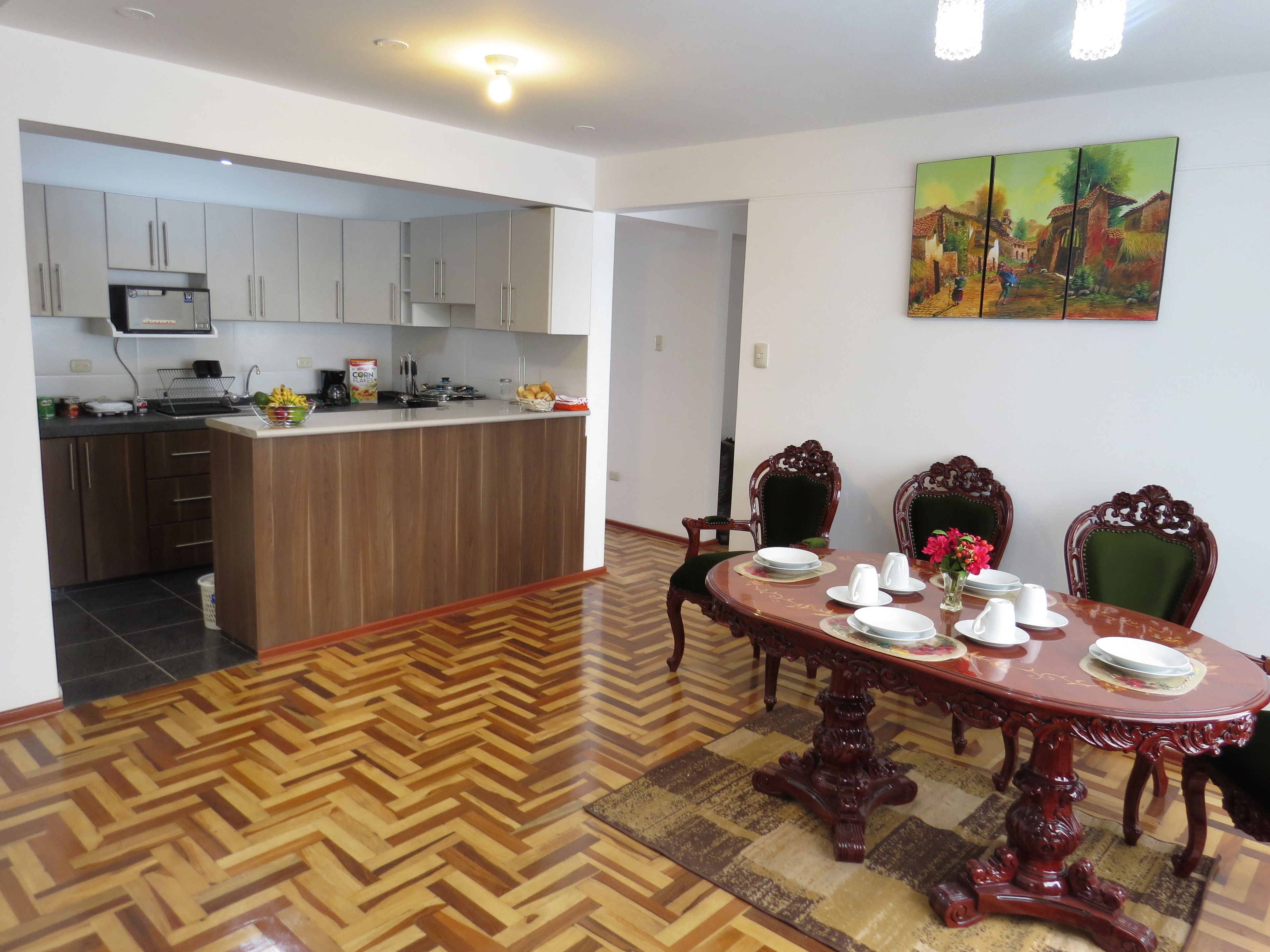 A dining area with a wooden table set for four, surrounded by ornate chairs, a kitchen with modern appliances, and a painting on the wall.