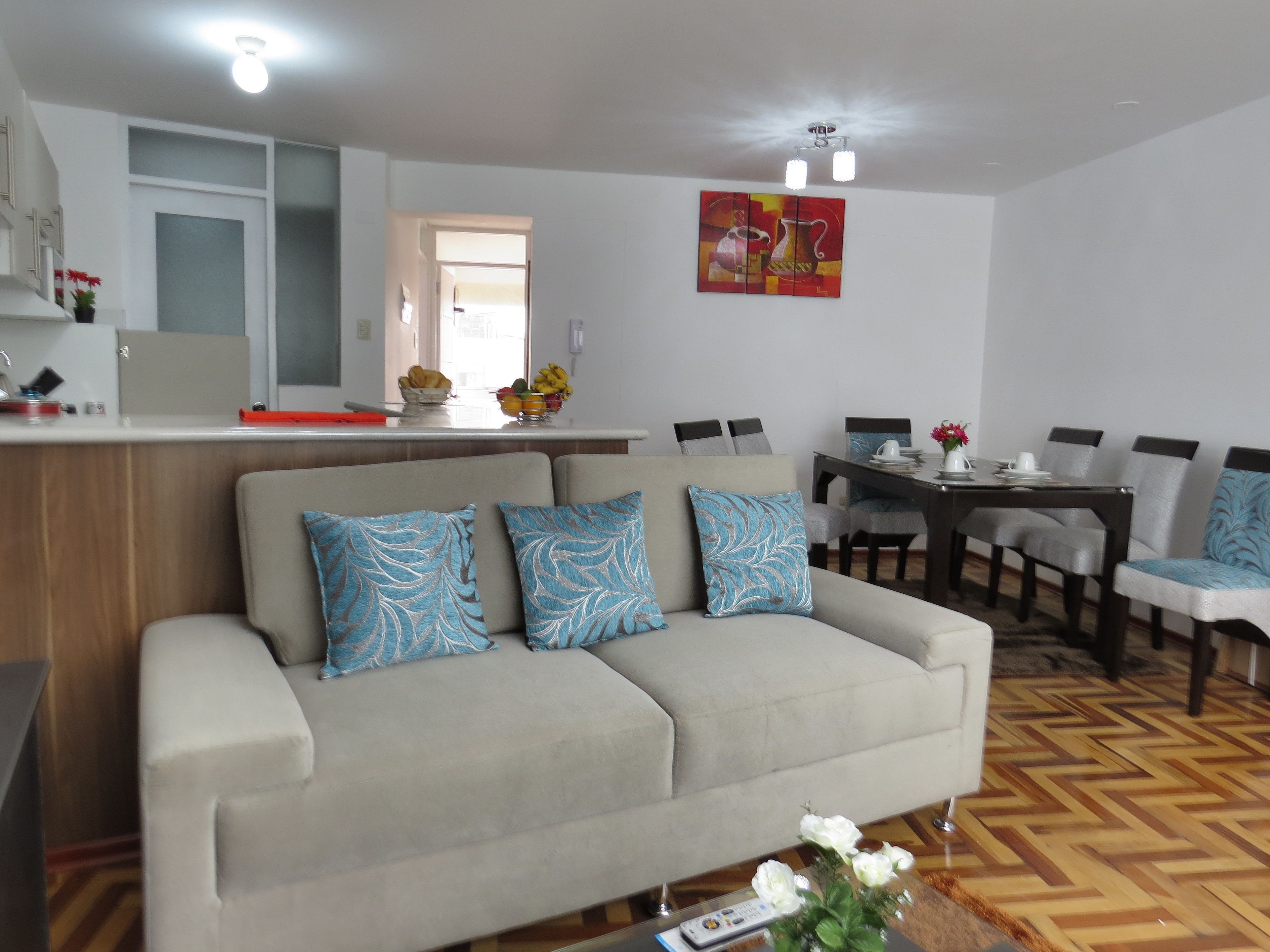 A living room with a sofa, a dining table with chairs, a kitchen area, and a painting on the wall.