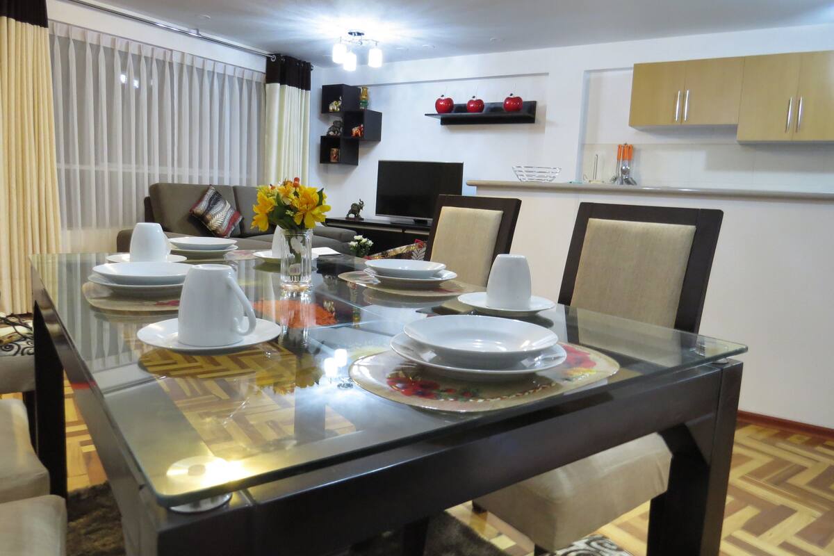 Un coin repas avec une table en verre, dressée pour quatre personnes, entourée de chaises. Un vase rempli de fleurs jaunes est posé sur la table.