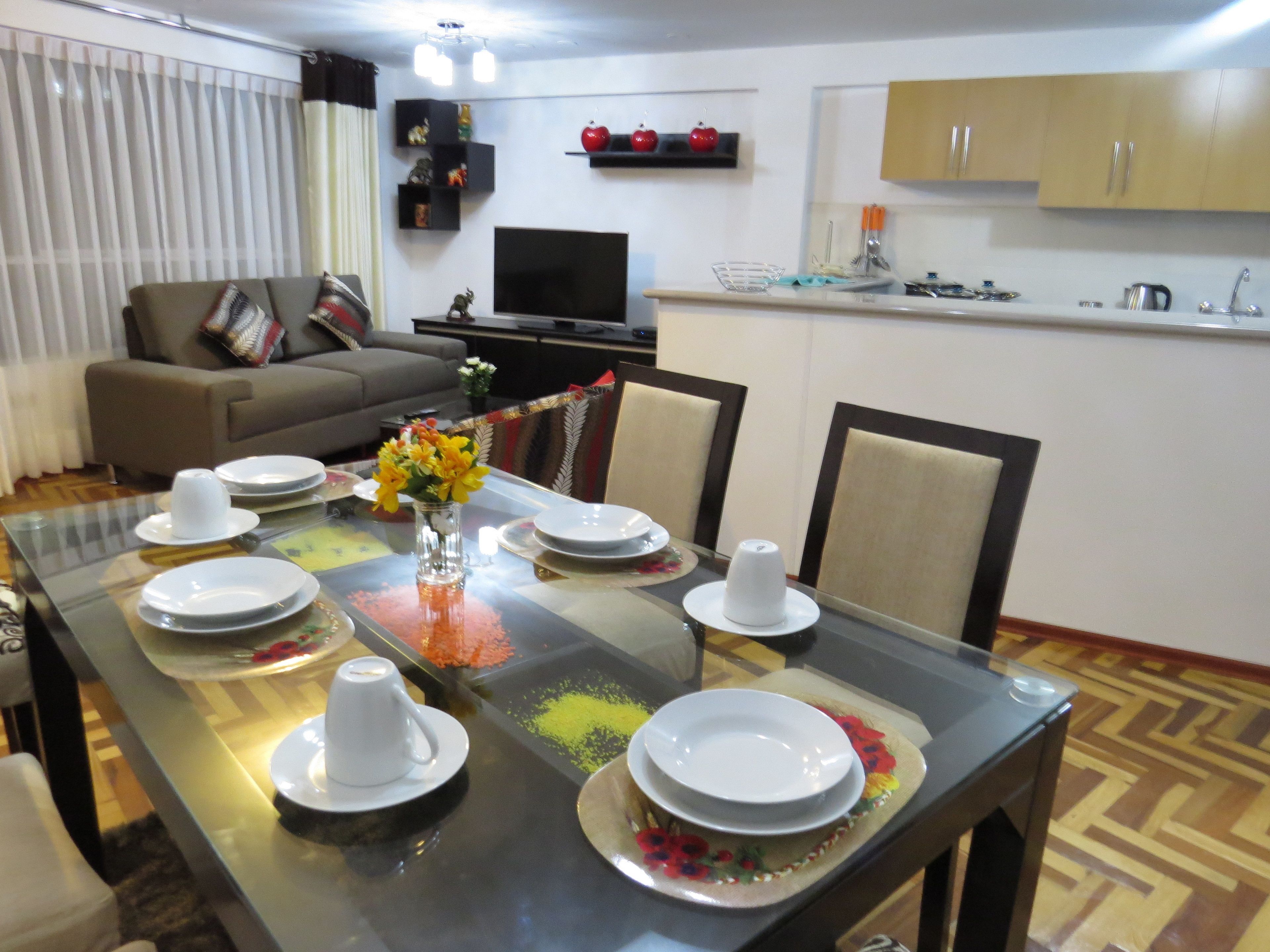 A dining area with a glass table set for four, surrounded by chairs. A sofa and a television are visible in the background.