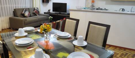 A dining area with a glass table set for four, surrounded by chairs. A sofa and a television are visible in the background.