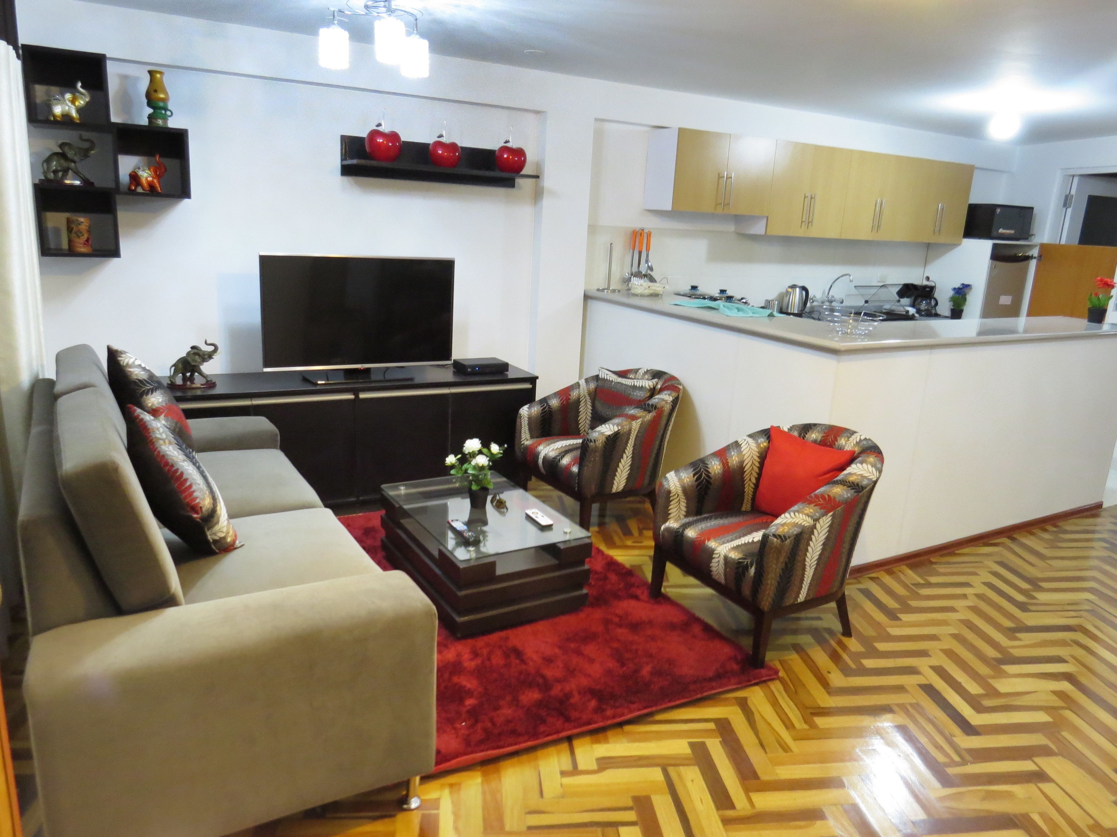 A living room with a sofa, armchairs, a coffee table, and a TV. There is a kitchen area with cabinets and a microwave.