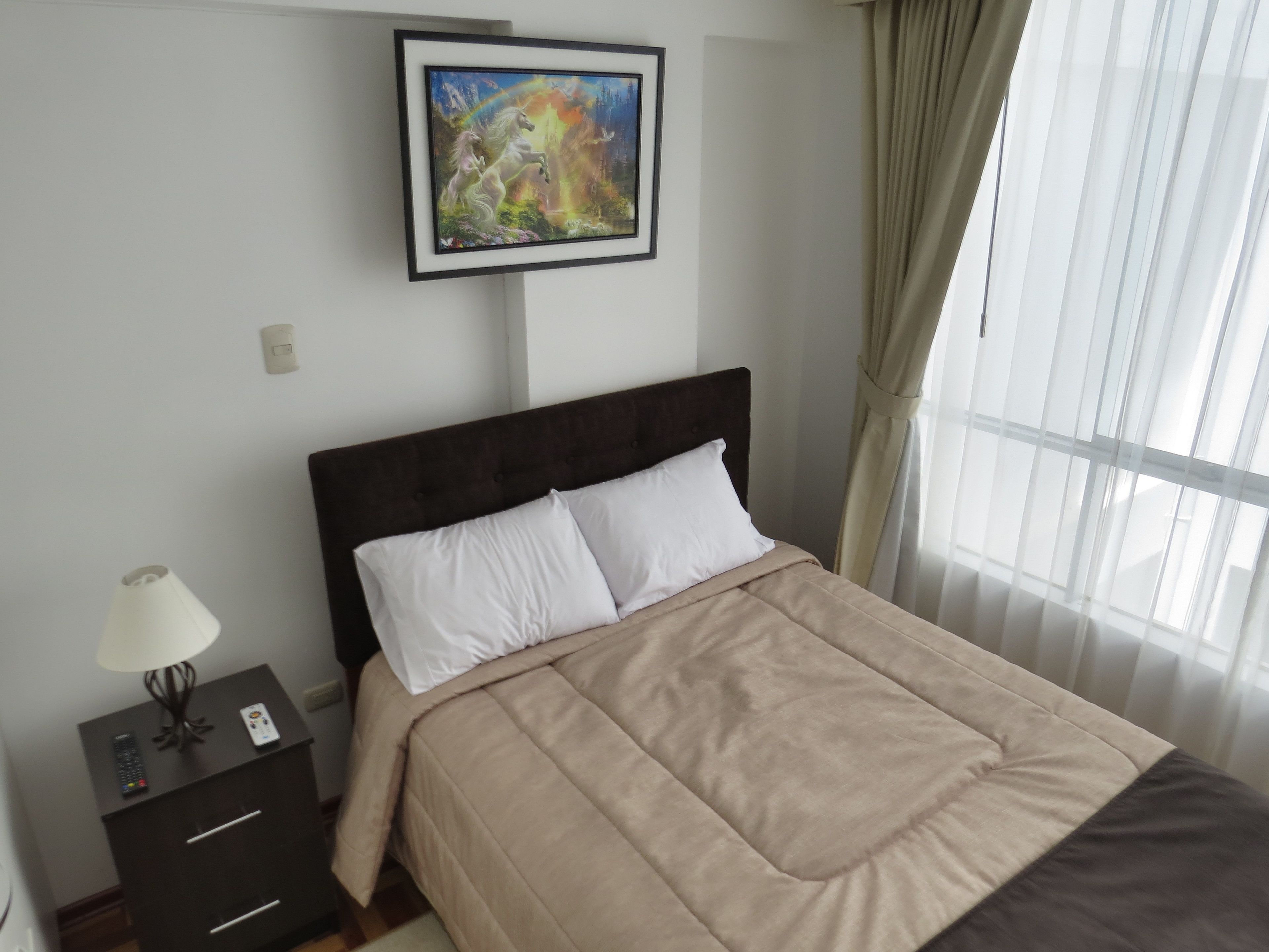 A neatly made bed with a headboard, a bedside table with a lamp, and a framed painting above the bed.