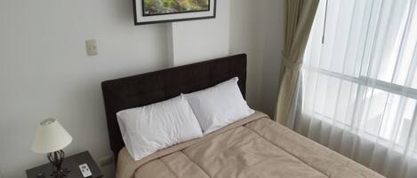 A neatly made bed with a headboard, a bedside table with a lamp, and a framed painting above the bed.