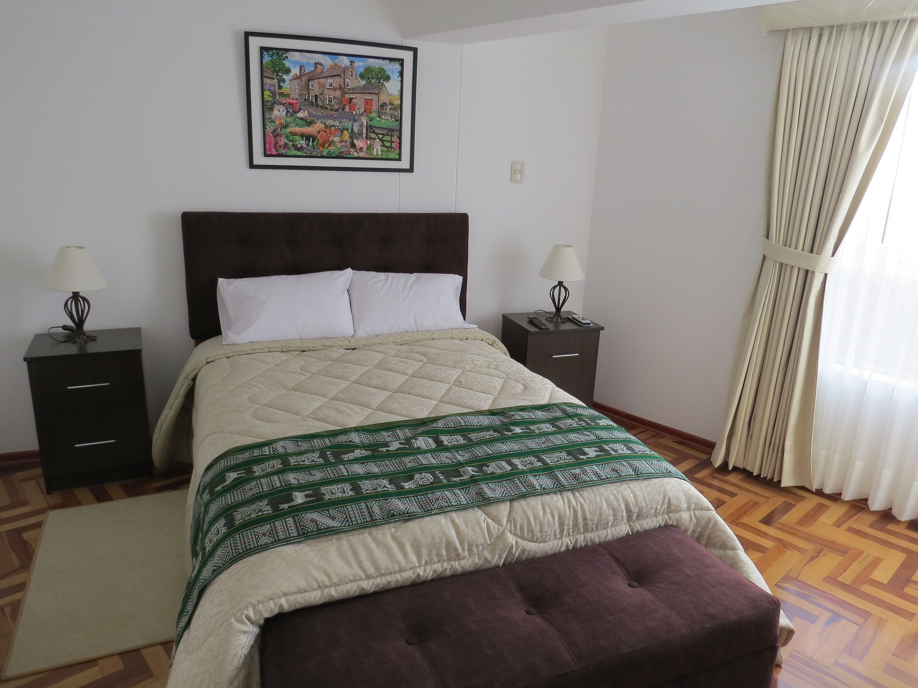 Quarto com cama, duas mesas de cabeceira, um criado-mudo, um quadro emoldurado e uma janela com cortinas.