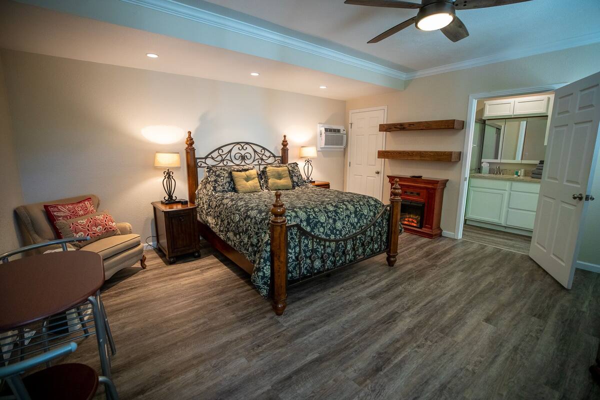 Quarto com cama de madeira, mesa de cabeceira, luminária, cadeira e mesa de jantar.