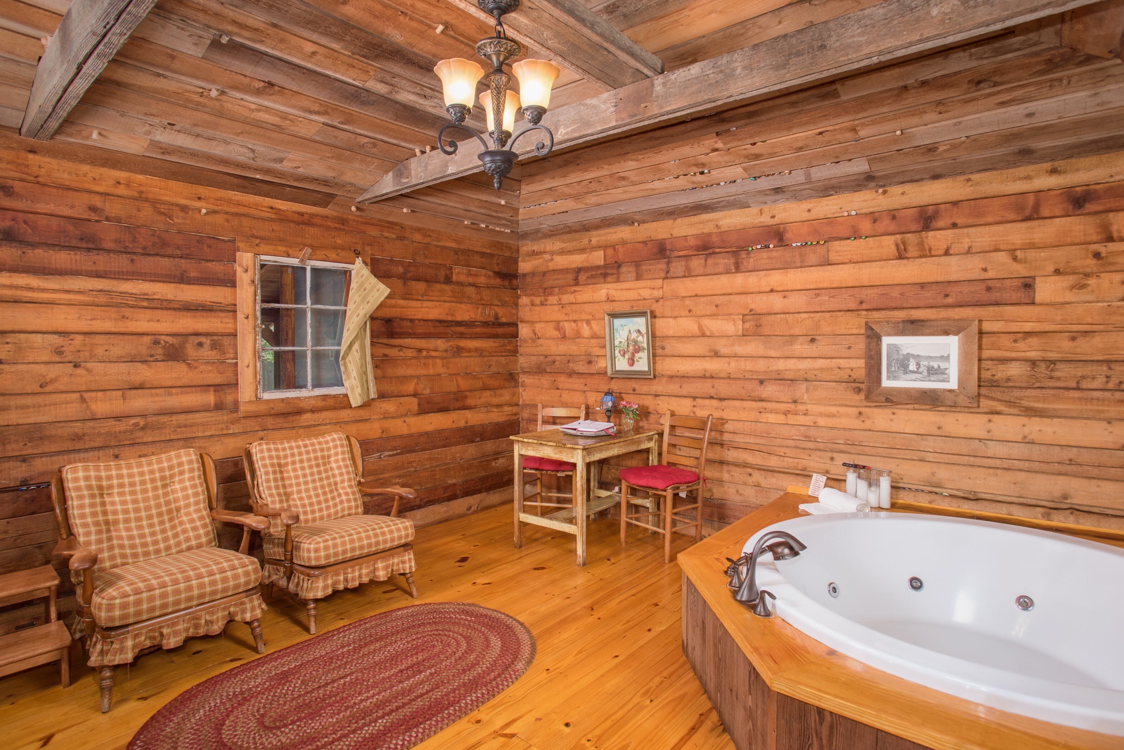 A wooden cabin interior with a wooden floor, two plaid chairs, a wooden table, a red rug, a large bathtub, and a ceiling fan.