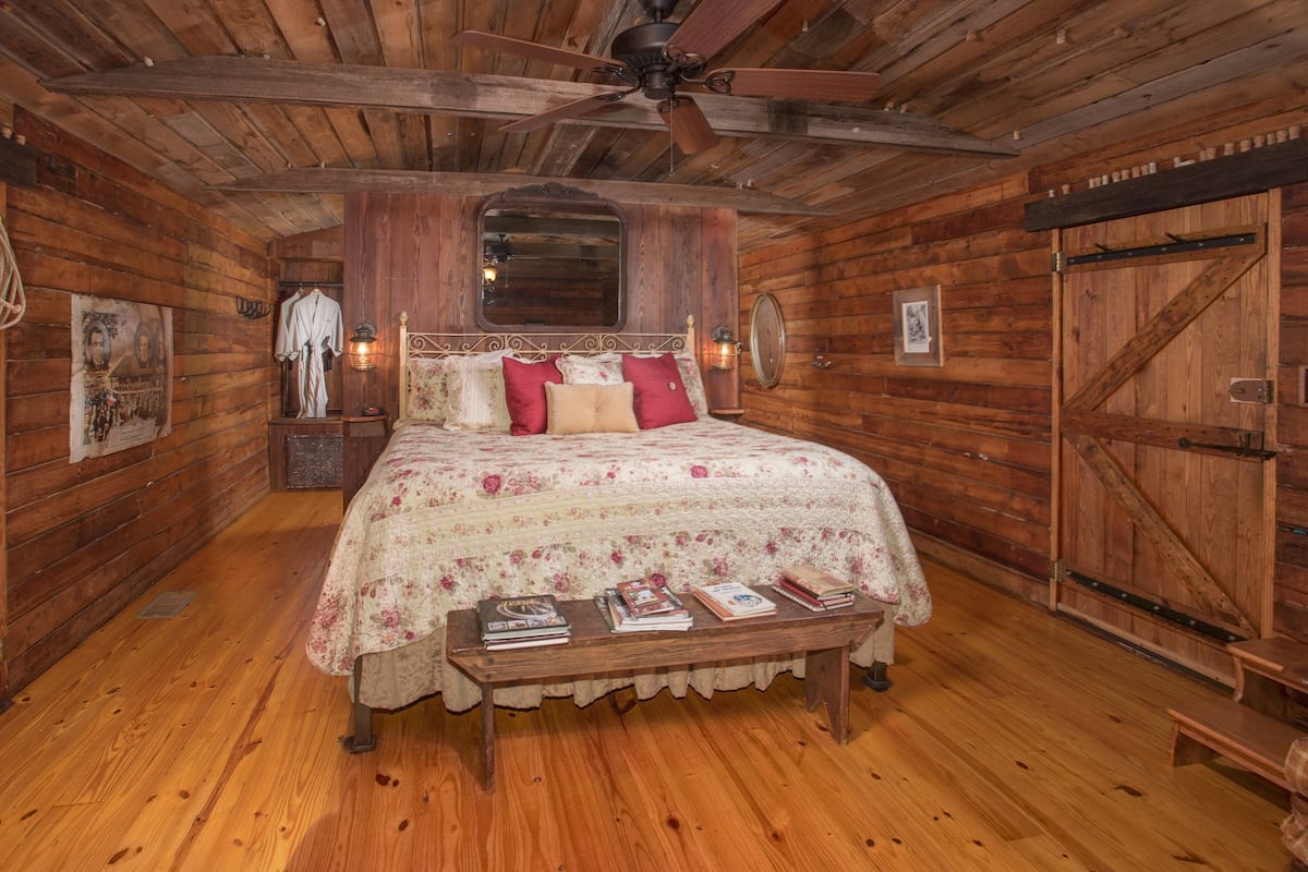 A wooden cabin bedroom with a bed, a ceiling fan, a wooden bench with books, and a large wooden door.