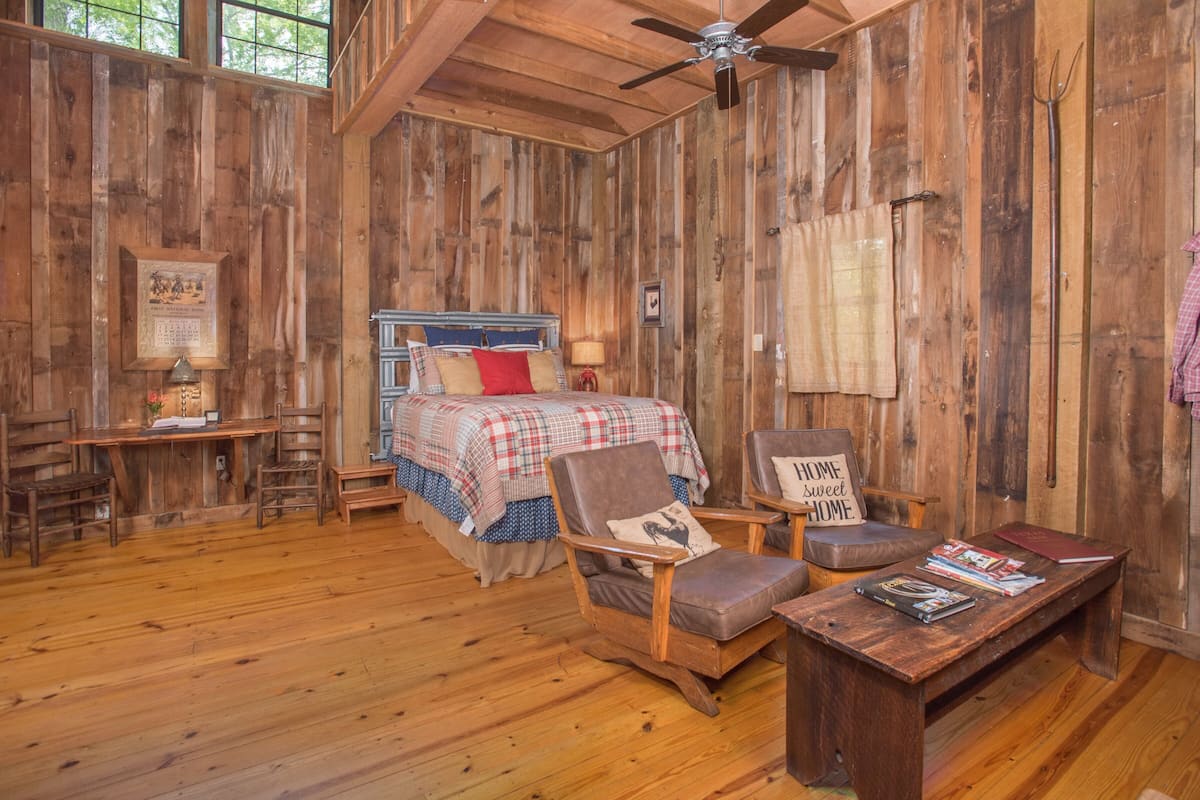 A rustic wooden room with a bed, two chairs, a desk, and a ceiling fan.
