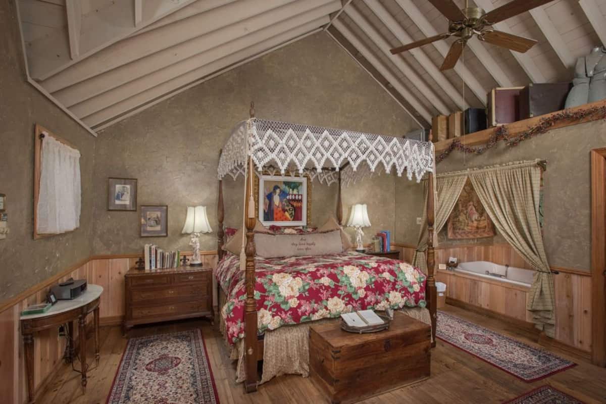 A bedroom with a four-poster bed, wooden furniture, a ceiling fan, and a bathtub.