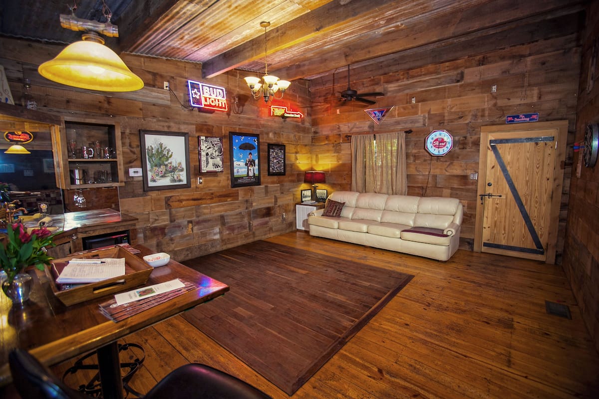 A cozy log cabin interior with a wooden floor, a white leather sofa, a dining table with a vase of flowers, and various framed pictures on the walls.