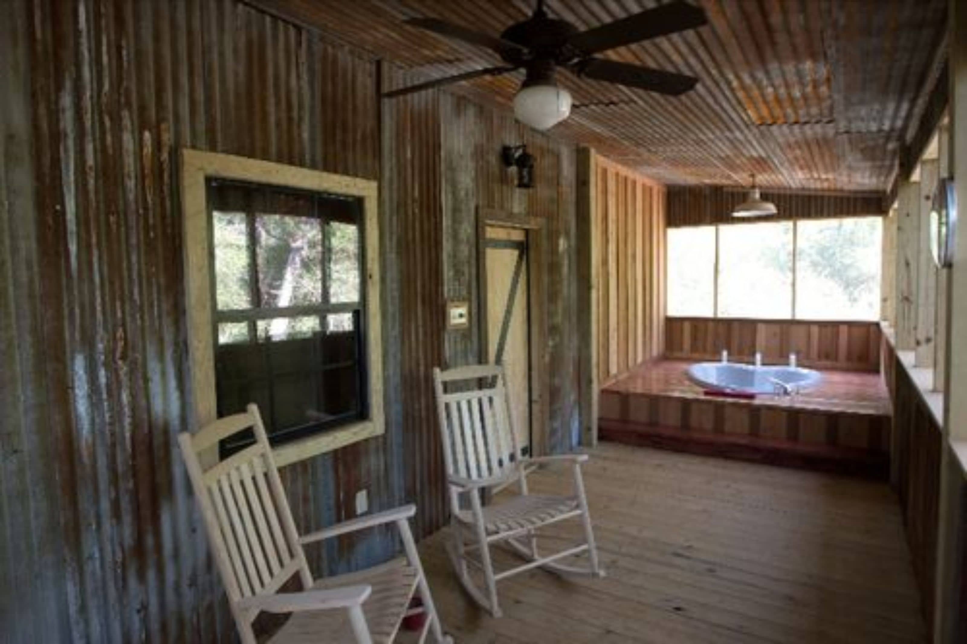 A rustic cabin with a wooden deck, two rocking chairs, a window, and a hot tub.