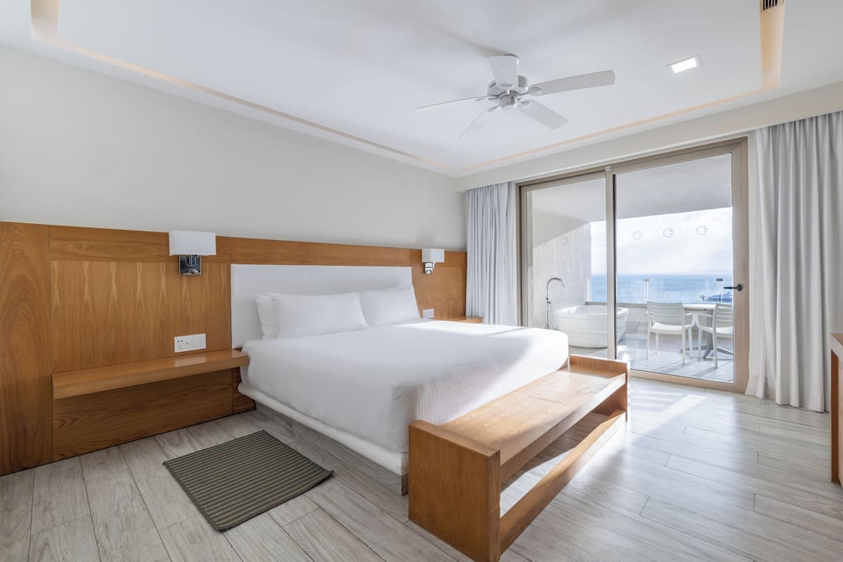 A modern hotel room with a large bed, wooden headboard, a ceiling fan, a sliding glass door to a balcony, and a small wooden bench.