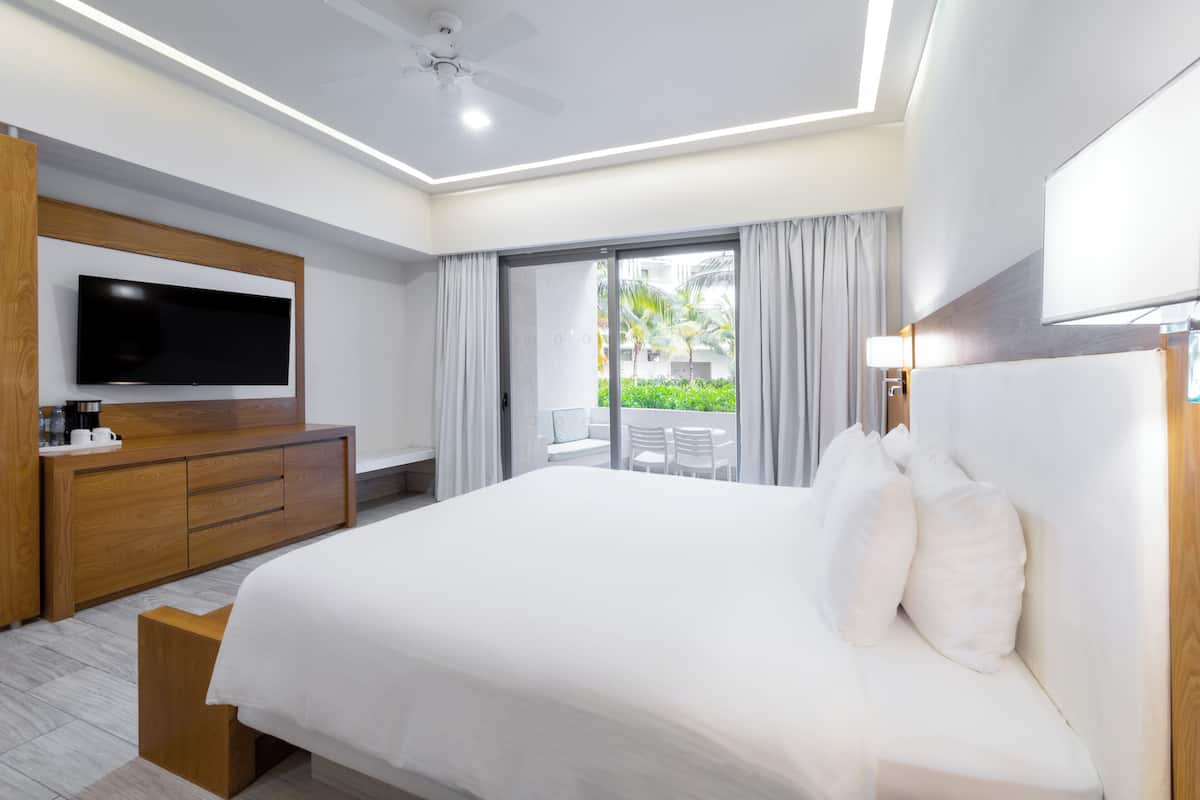 A modern hotel room with a large bed, a flat-screen TV, wooden furniture, and a balcony with outdoor seating.
