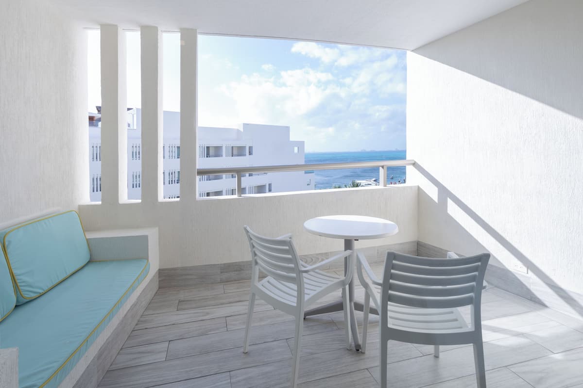 A balcony with a white sofa, two white chairs, a small round table, and a view of buildings and the sea.