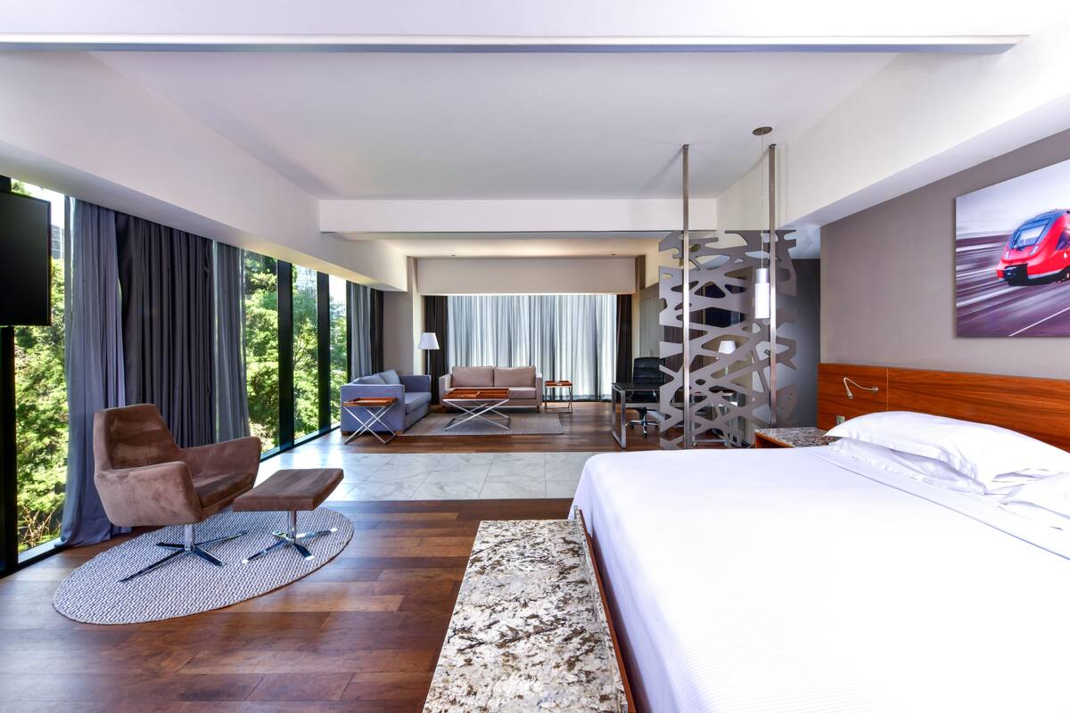 A modern hotel room with a large bed, a seating area with a sofa and armchairs, a desk, and a large window with a view of greenery.