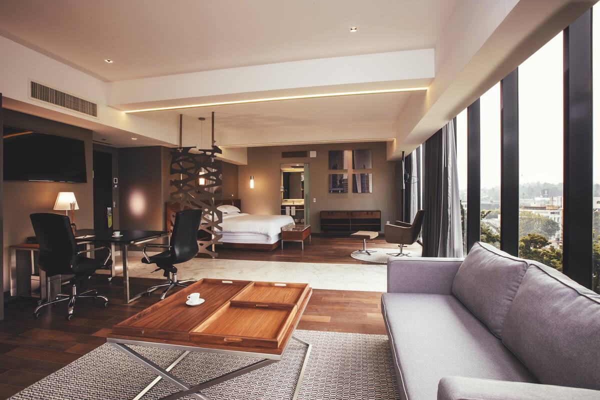 A modern hotel room with a large bed, a sofa, a desk with chairs, and a TV.
