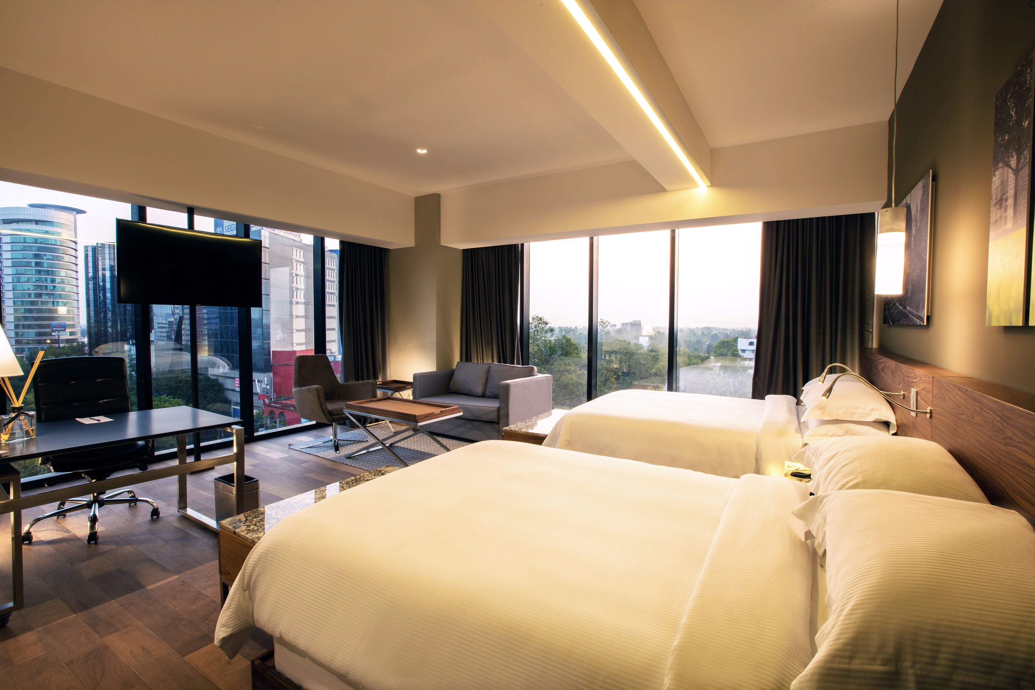 A hotel room with two beds, a desk with a computer, a TV, and large windows with a city view.