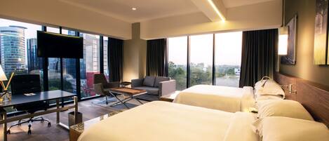 A hotel room with two beds, a desk with a computer, a TV, and large windows with a city view.