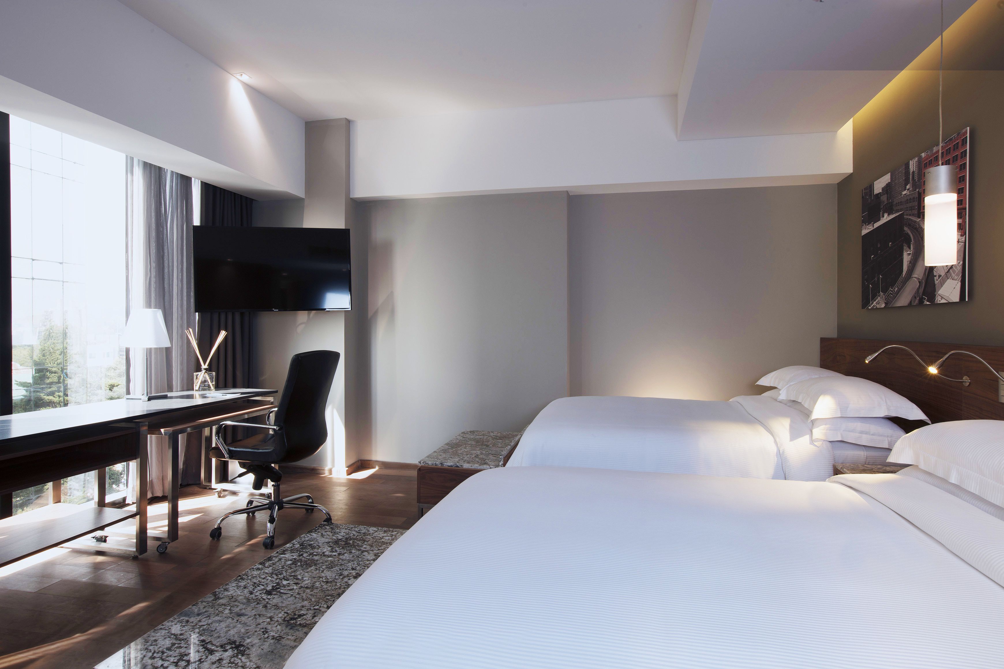Ein modernes Hotelzimmer mit einem großen Bett, einem Schreibtisch mit Stuhl, einem Flachbildfernseher und einem großen Fenster mit Blick auf die Stadt.