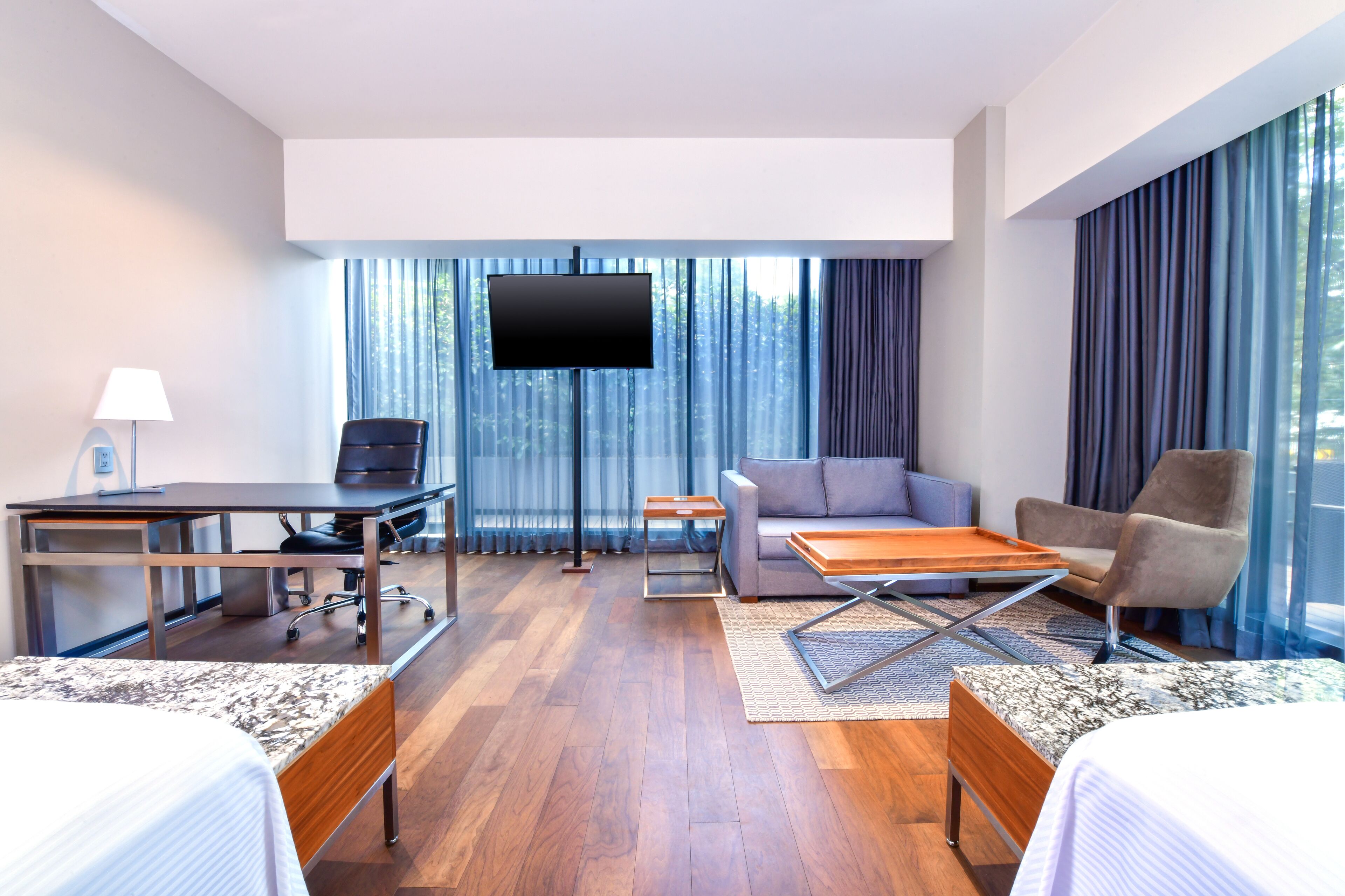 Quarto de hotel com cama, escrivaninha, sofá, televisão e janelas amplas com cortinas.