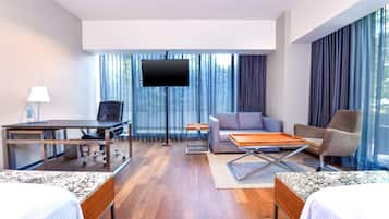 Quarto de hotel com cama, escrivaninha, sofá, televisão e janelas amplas com cortinas.