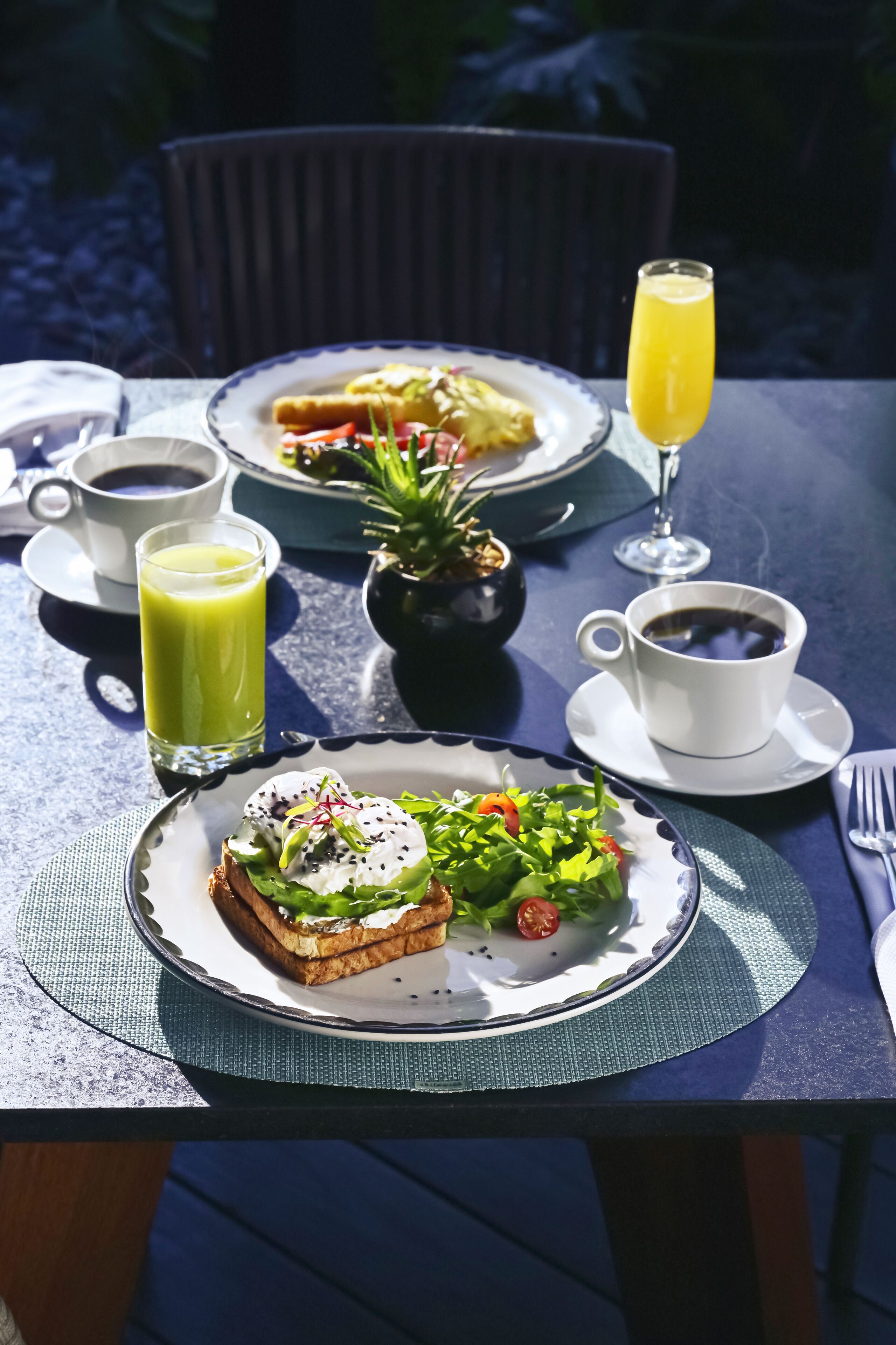 A table with breakfast items including toast, salad, and juice, with a chair and a small potted plant in the background.
