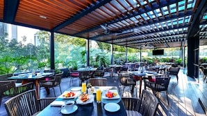 Un área de comedor al aire libre con mesas y sillas, rodeada de follaje y una terraza de madera.