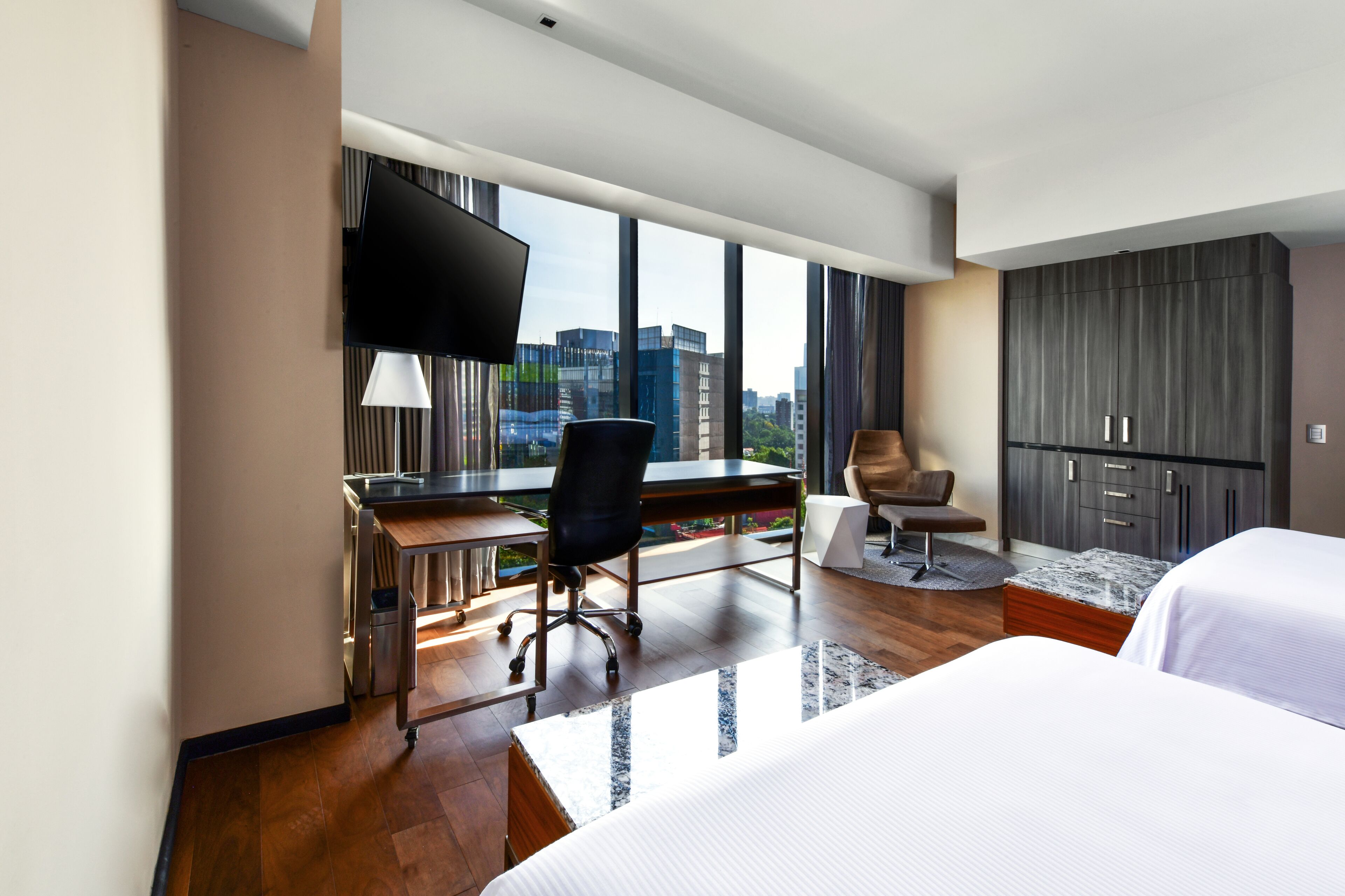 A hotel room with a large window, a desk with a chair, a bed, and a wardrobe.