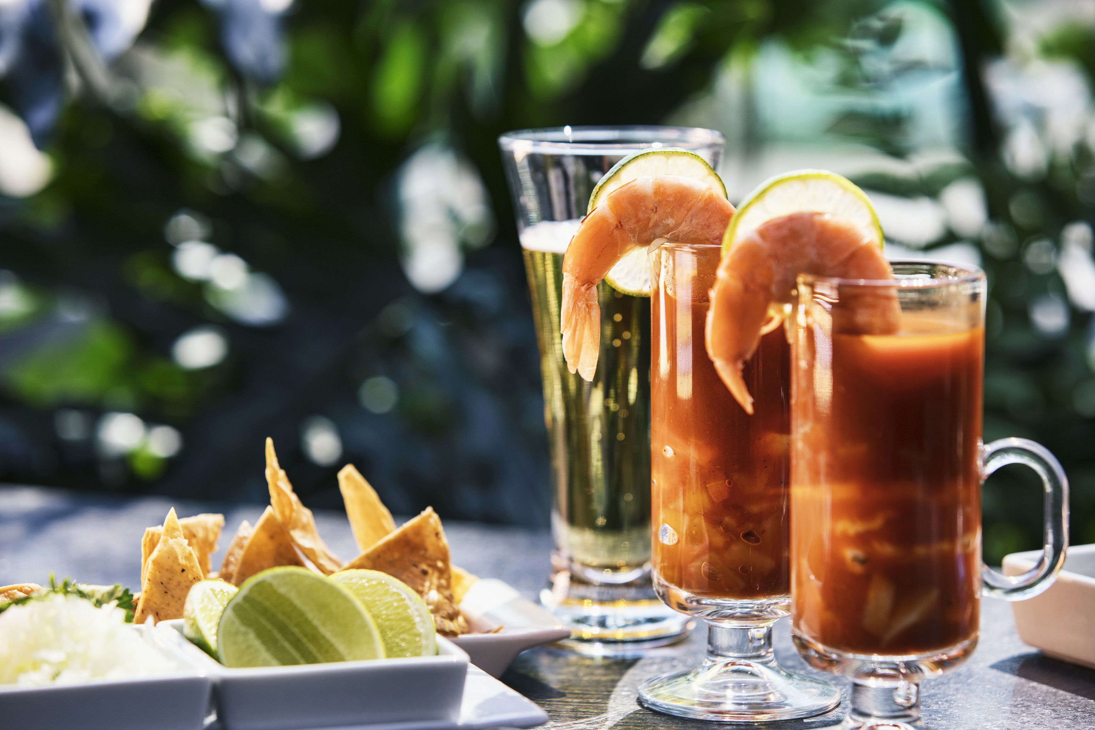 Two glasses of iced beverages with lime slices and a shrimp garnish, accompanied by tortilla chips and a small plate of white onions.