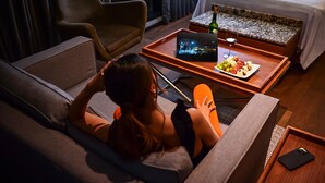 Una persona descansando en un sofá con una bandeja que contiene un ordenador portátil, una botella y aperitivos.