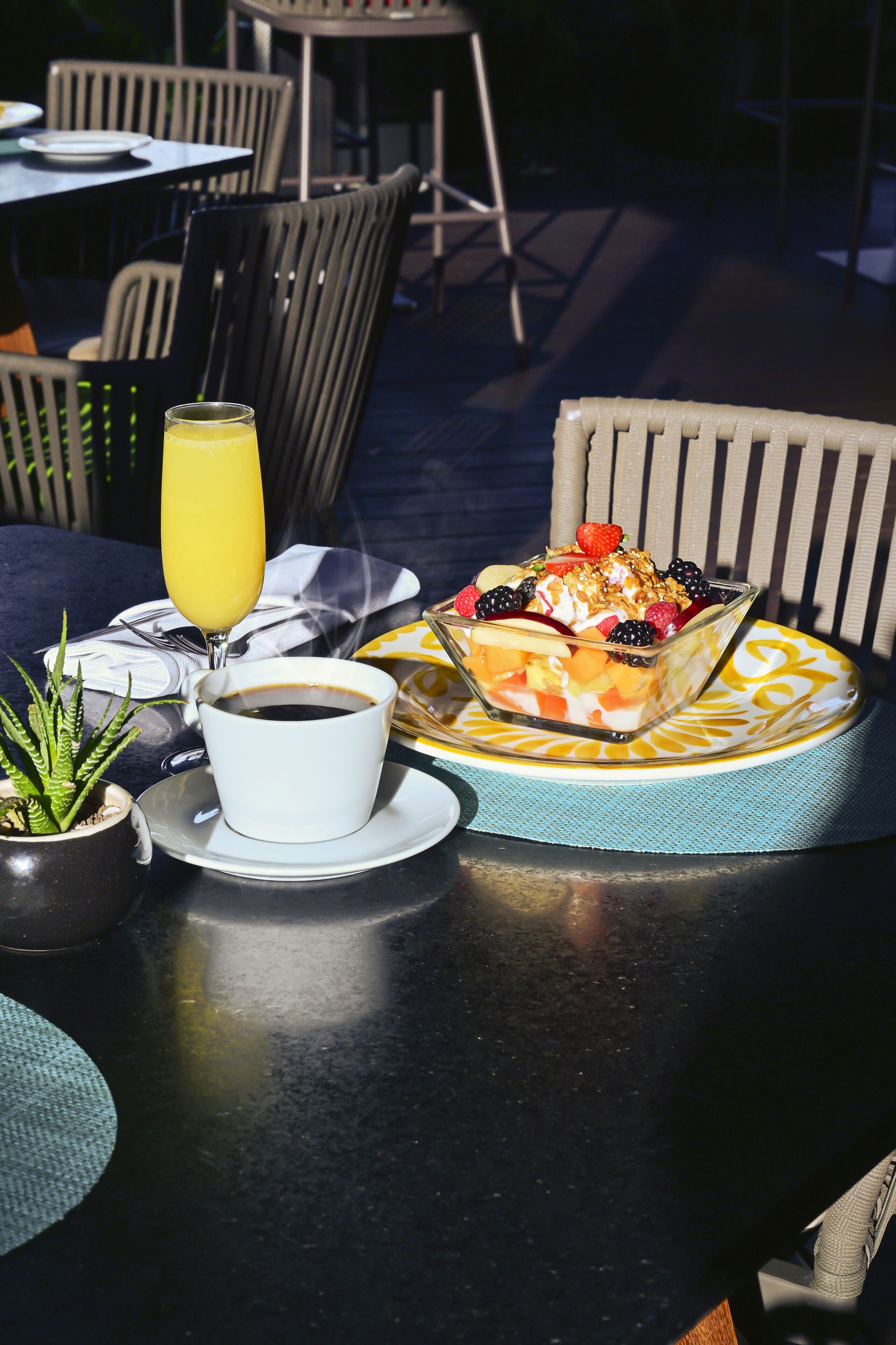 A table with a coffee cup, a glass of orange juice, and a fruit salad on a patterned plate.