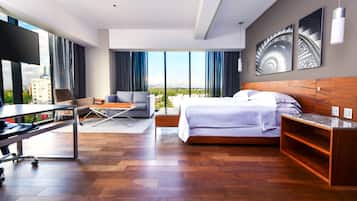 A modern hotel room with a large bed, a desk, and a chair. There is a large window with a view of the city.