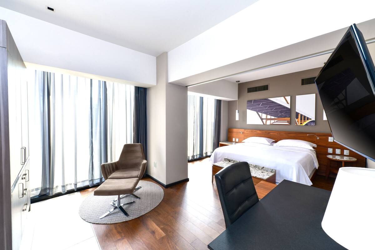 A hotel room with a bed, a desk, a chair, a TV, and large windows.