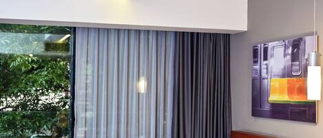A hotel room with two beds, a large window with curtains, a view of greenery outside, and a painting on the wall.