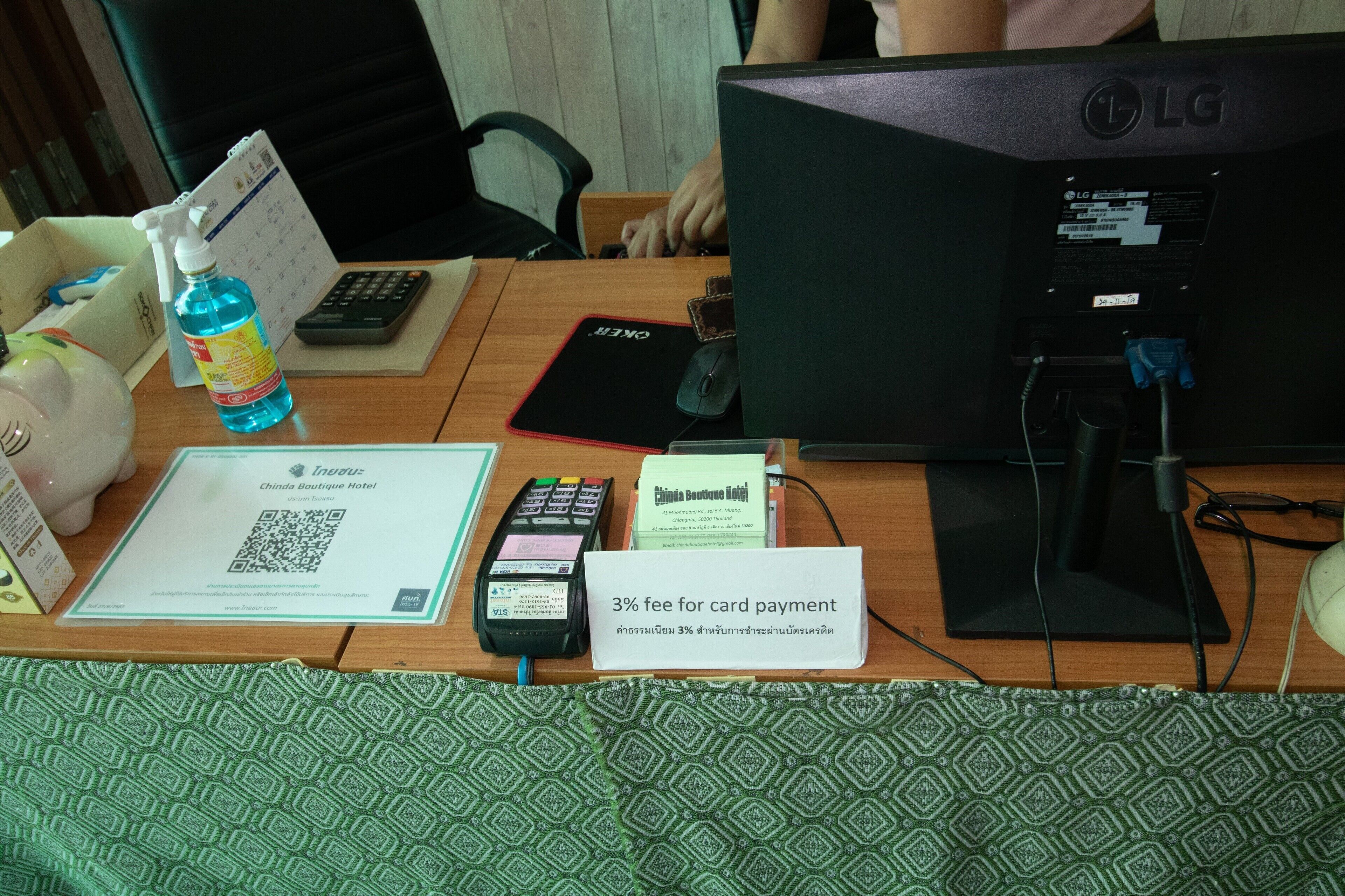A hotel desk with a computer, a card reader, a calendar, a bottle of hand sanitizer, and a sign indicating a 3% fee for card payments.