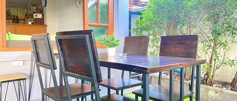 An outdoor dining area with a wooden table and chairs, a window with a view of greenery, and a small indoor seating area visible through the window.