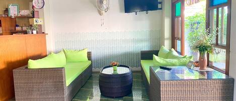 A room with wicker furniture, a flat-screen TV, a clock, and a window with a view of greenery.
