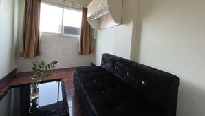 Un salón con sofá negro, una mesa de centro de vidrio y una planta en maceta. Hay una ventana con cortinas, un equipo de aire acondicionado y piso de baldosas.